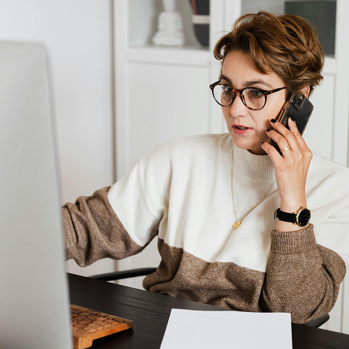Frau telefoniert am Handy während sie vor ihrem Computer arbeitet